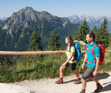 Das Bild zeigt einen Mann und eine Frau in kurzer Wanderkleidung in auf einem unbefestigten Weg laufen. Parallel zum Kiesweg verläuft ein Holzgeländern. Im Hintergrund erheben sich die Berge. Der Himmel ist blau. Die Sonne scheint.