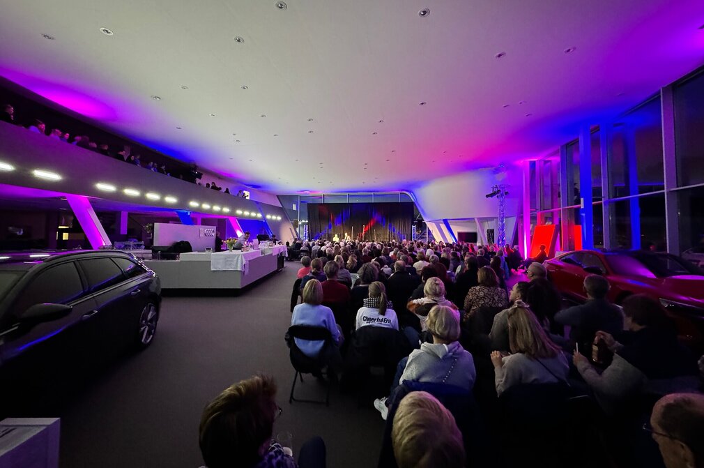 Das Bild zeigt mehrere Personen in Neoprenanzügen im knietiefen Wasser stehen. Alle tragen unterschiedliche Mützen und halten brennende Fackeln in den Händen. Es Dämmert. Es ist schon fast dunkel.  Auf der linken Seite des Bildes steht ein Auto. Auf einem Tresen sind Getränke und Gläser zu sehen. Im Hintergrund sieht man eine Bühne mit mehreren Musikern darauf.