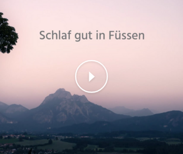 Das Bild zeigt eine in Abendlicht getauchte Landschaft mit einer imposanten Bergkulisse im Hintergrund. Der Himmel leuchtet in Lila- und Blautönen des letzten Sonnenlichts. Im Vordergrund rahmen zwei große Laubbäume das Bild links und rechts ein.