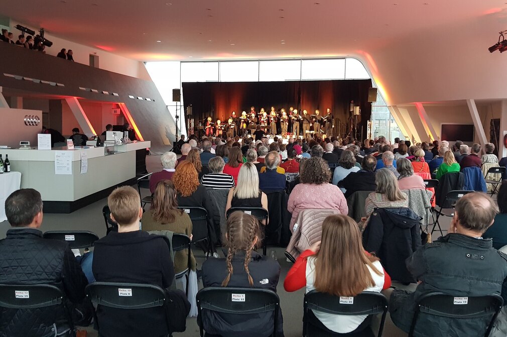 Chormomente Das Bild zeigt einen Chor auf einer Bühne stehen. Vor der Bühne sind zahlreiche Stuhlreihen zu sehen. Beinahe alle Plätze sind gefüllt. Naben den Stuhlreihen sind zwei Tresen aufgebaut, an denen Getränke und Gläser stehen. Der Raum ist hell erleuchtet.