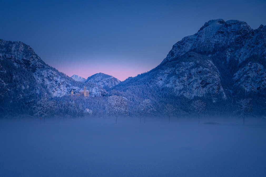An einem Wintermorgen mit Neuschnee ragt Schloss Neuschwanstein über einem nebligen Feld, umringt von Bergen auf. 