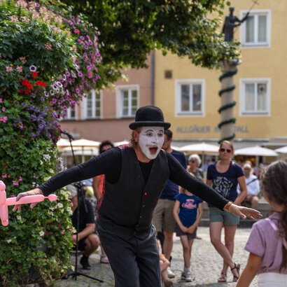 Das Bild zeigt einen Pantomimen, der ein Luftballontier in der rechten Hand hält. Er ist schwarz gekleidet, trägt einen schwarzen Hut und ist weiß geschminkt. Er steht in einer Fußgängerzone. Mehrere Personen sind zu sehen. Im Hintergrund sieht man bunte Hausfassaden. Die Sonne scheint. 