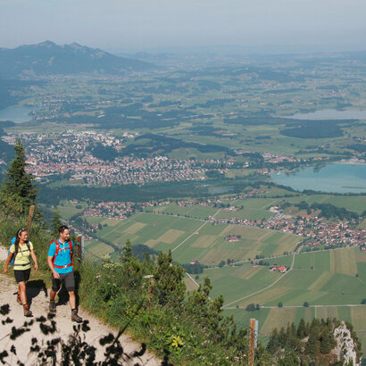 Ein Paar wandert auf dem Gipfel des Tegelbergs. Im Hintergrund sind Füssen, der Forggensee und die Tannheimer Alpen zu sehen