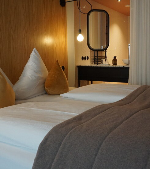  Das Bild zeigt ein modernes Hotelzimmer, das ein Gefühl von Luxus und Komfort vermittelt. Dominierend ist das große Bett, das in weiße Leintücher gehüllt ist und mit zwei goldfarbenen Kissen geschmückt ist. Das Bett steht an einer hölzernen Wand, die dem Raum eine warme Note verleiht.