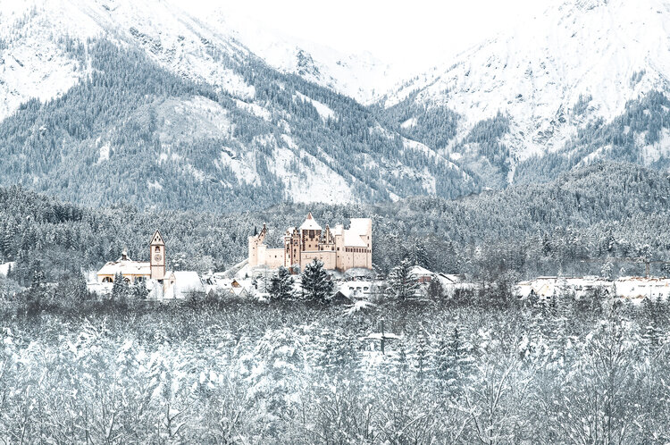 Blick von einem Hügel auf Altstadt von Füssen. Prominent zu sehen sind das Hohe Schloss und die Kirche vor schneebedeckten Wäldern und Bergen.
