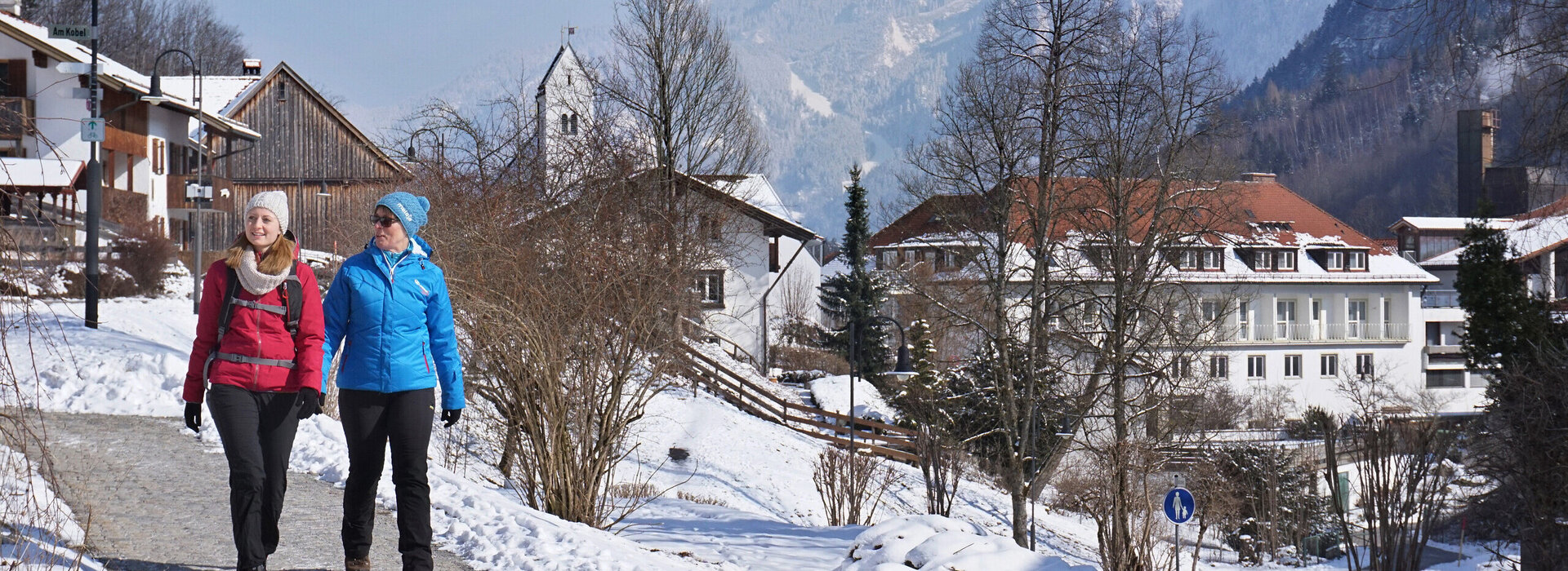 Auf einer Straße laufen zwei Frauen, die winterlich gekleidet sind. Es sind Häuser und auch ein Kirchturm zu sehen. Der Boden und die Dächer der Gebäude sind schneebedeckt. Im Hintergrund erheben sich die ebenfalls schneebedeckten Berge. Der Himmel ist blau. Die Sonne scheint. 