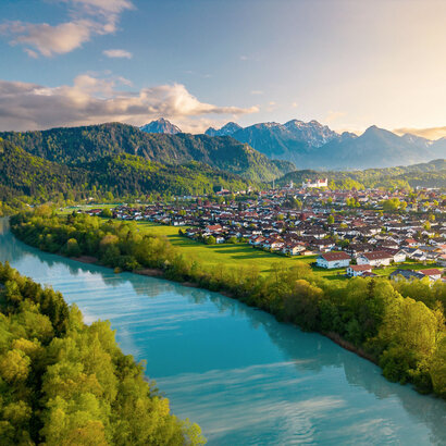 Das Bild zeigt einen breiten türkisblauen Fluss, der ruhig vor sich hinfließt. Am Ufer des Flusses wachsen Bäume. Auf der linken Uferseite beginnt der Bergwald. Am rechten Ufer erstreckt sich eine Wiese, die an die Stadt Füssen grenzt. Über Füssen thront auf einer Anhöhe das Hohe Schloss. Im Hintergrund ragen, teilweise schneebedeckte Berge empor. Die die Stadt und den Fluss einrahmen.  
