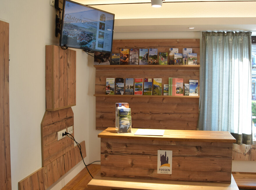 Das Bild zeigt ein Detail eines Raumes. Vor einer Holzeckbank steht ein hölzerner Tresen. An der Wand dahinter ist ein Prospektregal angebracht, das mit zahleichen Prospekten gefüllt ist. An der Nebenwand des Tresens ist ein Monitor angebracht. 