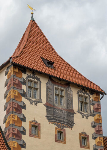 Das Bild zeigt ein großes, reich verziertes Gebäude mit einem charakteristischen Architekturstil. Das Gebäude ist in einer Vielzahl von Farben gestaltet, darunter Töne von Gelb, Orange und Braun. Das Dach des Gebäudes ist rot und weist eine spitze, konische Form auf. Auf dem Dach ist eine kleine gelbe Fahne. Das Gebäude hat mehrere Fenster, die alle mit filigranen Designs und Mustern verziert sind. Der Hintergrund ist wolkig, was auf einen bedeckten Tag hindeutet. Der Eindruck ist der eines großen, historischen Gebäudes, möglicherweise eines Schlosses oder einer Kirche, mit einem reichen und detaillierten architektonischen Design.