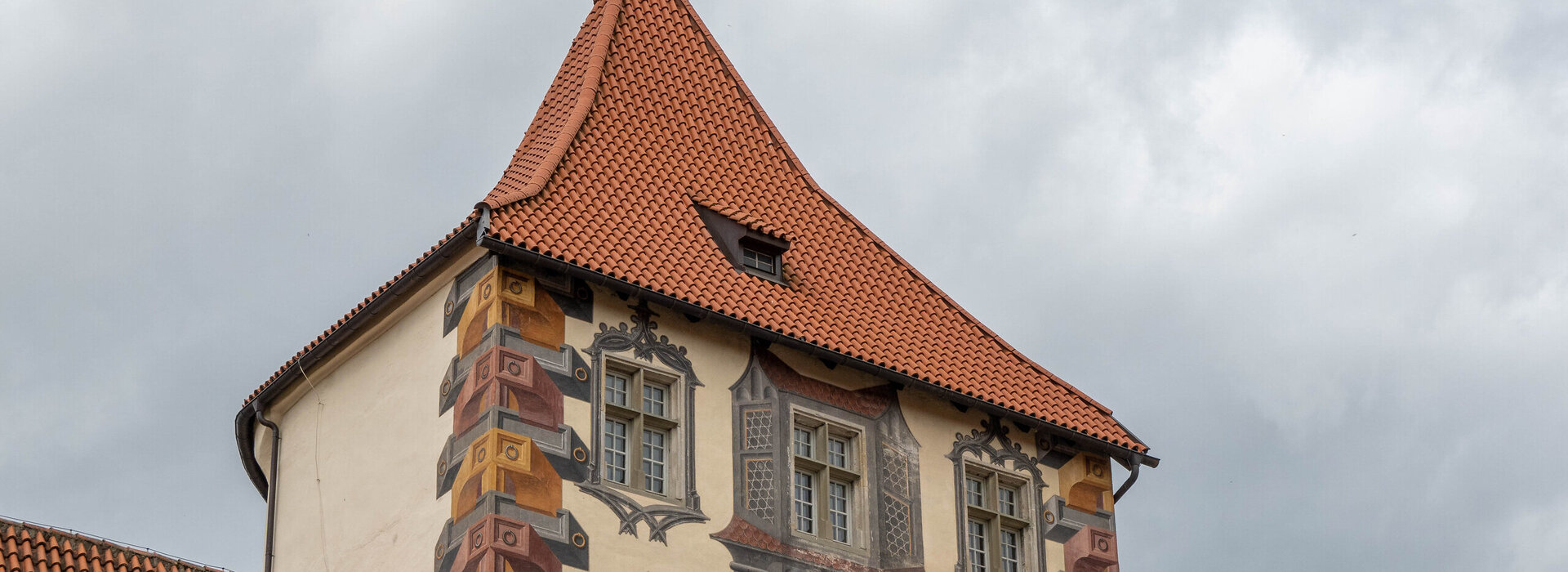 Das Bild zeigt ein großes, reich verziertes Gebäude mit einem charakteristischen Architekturstil. Das Gebäude ist in einer Vielzahl von Farben gestaltet, darunter Töne von Gelb, Orange und Braun. Das Dach des Gebäudes ist rot und weist eine spitze, konische Form auf. Auf dem Dach ist eine kleine gelbe Fahne. Das Gebäude hat mehrere Fenster, die alle mit filigranen Designs und Mustern verziert sind. Der Hintergrund ist wolkig, was auf einen bedeckten Tag hindeutet. Der Eindruck ist der eines großen, historischen Gebäudes, möglicherweise eines Schlosses oder einer Kirche, mit einem reichen und detaillierten architektonischen Design.