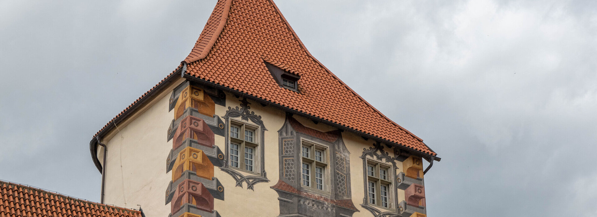 Das Bild zeigt ein großes, reich verziertes Gebäude mit einem charakteristischen Architekturstil. Das Gebäude ist in einer Vielzahl von Farben gestaltet, darunter Töne von Gelb, Orange und Braun. Das Dach des Gebäudes ist rot und weist eine spitze, konische Form auf. Auf dem Dach ist eine kleine gelbe Fahne. Das Gebäude hat mehrere Fenster, die alle mit filigranen Designs und Mustern verziert sind. Der Hintergrund ist wolkig, was auf einen bedeckten Tag hindeutet. Der Eindruck ist der eines großen, historischen Gebäudes, möglicherweise eines Schlosses oder einer Kirche, mit einem reichen und detaillierten architektonischen Design.