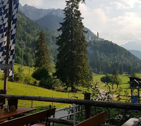 Das Bild zeigt im Vordergrund ein paar Biertische und Bänke und eine wehende Bayerische Fahne. Im Hintergrund erheben sich hinter grünen Wiesen mächtige Berge und das Märchenschloss Neuschwanstein.