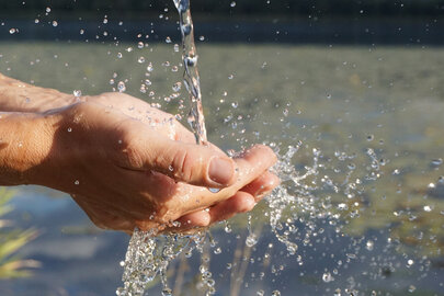 Das Bild fängt einen Moment der Ruhe in der Natur ein. Eine menschliche Hand ist im Vordergrund zu sehen, auf die ein Wasserstrahl fällt. Von der Hand fließt das Wasser weiter in den sich darunter befindenden Hopfensee. Die Wassertröpfchen, die von der Hand wegspritzen sind in der Luft eingefroren, was eine dynamische und fesselnde Szene schafft. Im Hintergrund erstreckt sich eine majestätische Gebirgskette über den Horizont. Die Berge sind teilweise von einer Wolkenschicht verhüllt, was der Szene Tiefe und Geheimnis verleiht. Die Farben im Bild sind hauptsächlich blau und grün, was die natürlichen Elemente in der Szene widerspiegelt. Die Gesamtanordnung des Bildes suggeriert eine friedliche und ruhige Umgebung, in der die menschliche Hand und die natürlichen Elemente harmonisch koexistieren. Das Bild ist eine schöne Darstellung des Zusammenspiels zwischen Mensch und Natur.