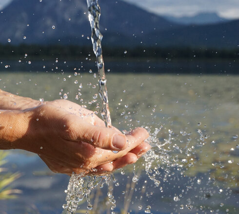 Das Bild fängt einen Moment der Ruhe in der Natur ein. Eine menschliche Hand ist im Vordergrund zu sehen, auf die ein Wasserstrahl fällt. Von der Hand fließt das Wasser weiter in den sich darunter befindenden Hopfensee. Die Wassertröpfchen, die von der Hand wegspritzen sind in der Luft eingefroren, was eine dynamische und fesselnde Szene schafft. Im Hintergrund erstreckt sich eine majestätische Gebirgskette über den Horizont. Die Berge sind teilweise von einer Wolkenschicht verhüllt, was der Szene Tiefe und Geheimnis verleiht. Die Farben im Bild sind hauptsächlich blau und grün, was die natürlichen Elemente in der Szene widerspiegelt. Die Gesamtanordnung des Bildes suggeriert eine friedliche und ruhige Umgebung, in der die menschliche Hand und die natürlichen Elemente harmonisch koexistieren. Das Bild ist eine schöne Darstellung des Zusammenspiels zwischen Mensch und Natur.