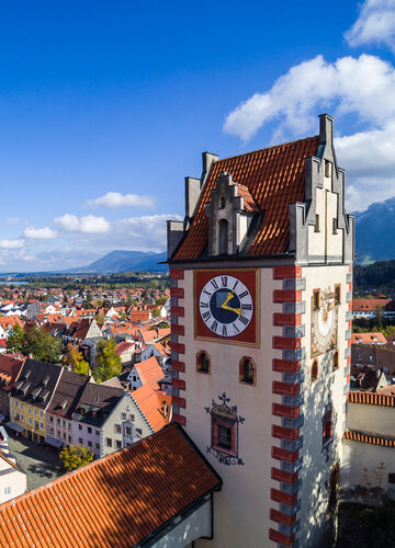 Das Foto zeigt Füssen von oben. Den Blick des Betrachters wird über die Dächer von Füssen geleitet. Im Vordergrund erhebt sich der sogenannte Uhrturm des Hohen Schlosses. Dahinter beginnen die Dächer der Füssener Altstadt. Im Hintergrund kann man schon die Berge und den türkisblauen Forggensee erkennen. 