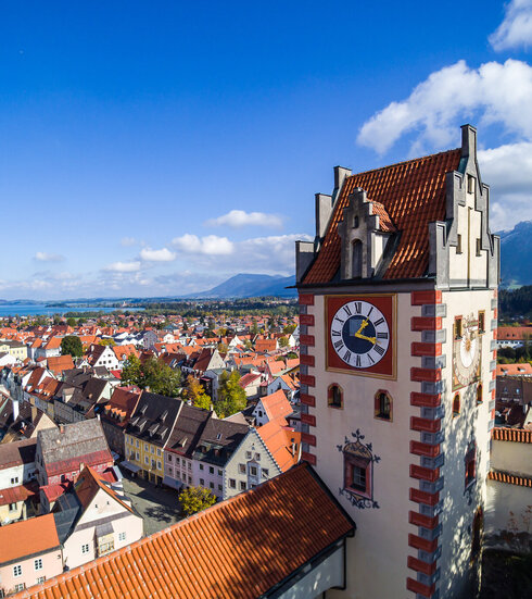 Das Foto zeigt Füssen von oben. Den Blick des Betrachters wird über die Dächer von Füssen geleitet. Im Vordergrund erhebt sich der sogenannte Uhrturm des Hohen Schlosses. Dahinter beginnen die Dächer der Füssener Altstadt. Im Hintergrund kann man schon die Berge und den türkisblauen Forggensee erkennen. 