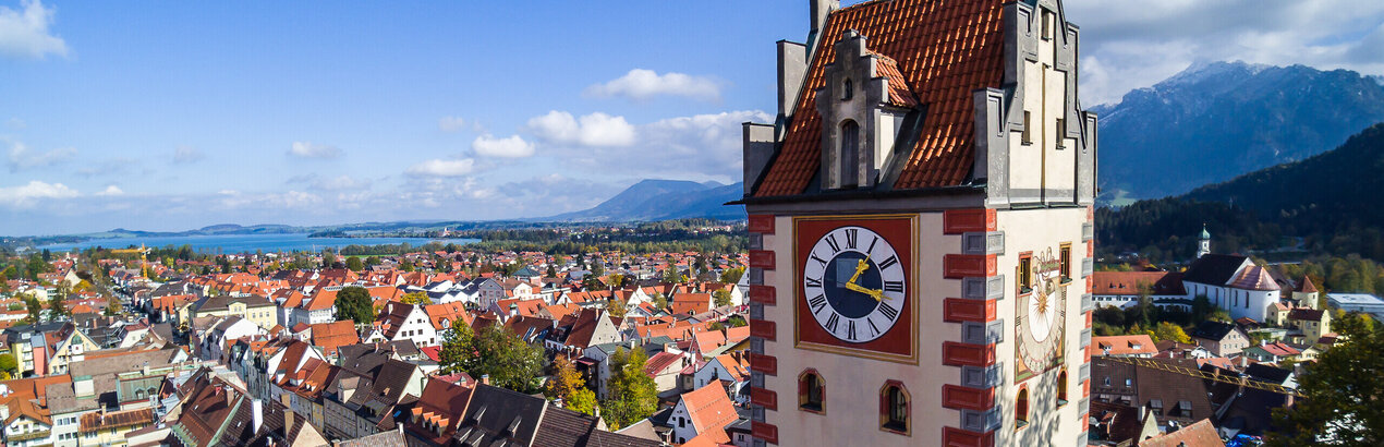 Das Foto zeigt Füssen von oben. Den Blick des Betrachters wird über die Dächer von Füssen geleitet. Im Vordergrund erhebt sich der sogenannte Uhrturm des Hohen Schlosses. Dahinter beginnen die Dächer der Füssener Altstadt. Im Hintergrund kann man schon die Berge und den türkisblauen Forggensee erkennen. 