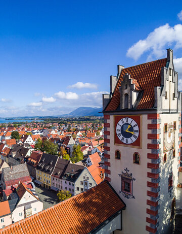 Das Foto zeigt Füssen von oben. Den Blick des Betrachters wird über die Dächer von Füssen geleitet. Im Vordergrund erhebt sich der sogenannte Uhrturm des Hohen Schlosses. Dahinter beginnen die Dächer der Füssener Altstadt. Im Hintergrund kann man schon die Berge und den türkisblauen Forggensee erkennen. 