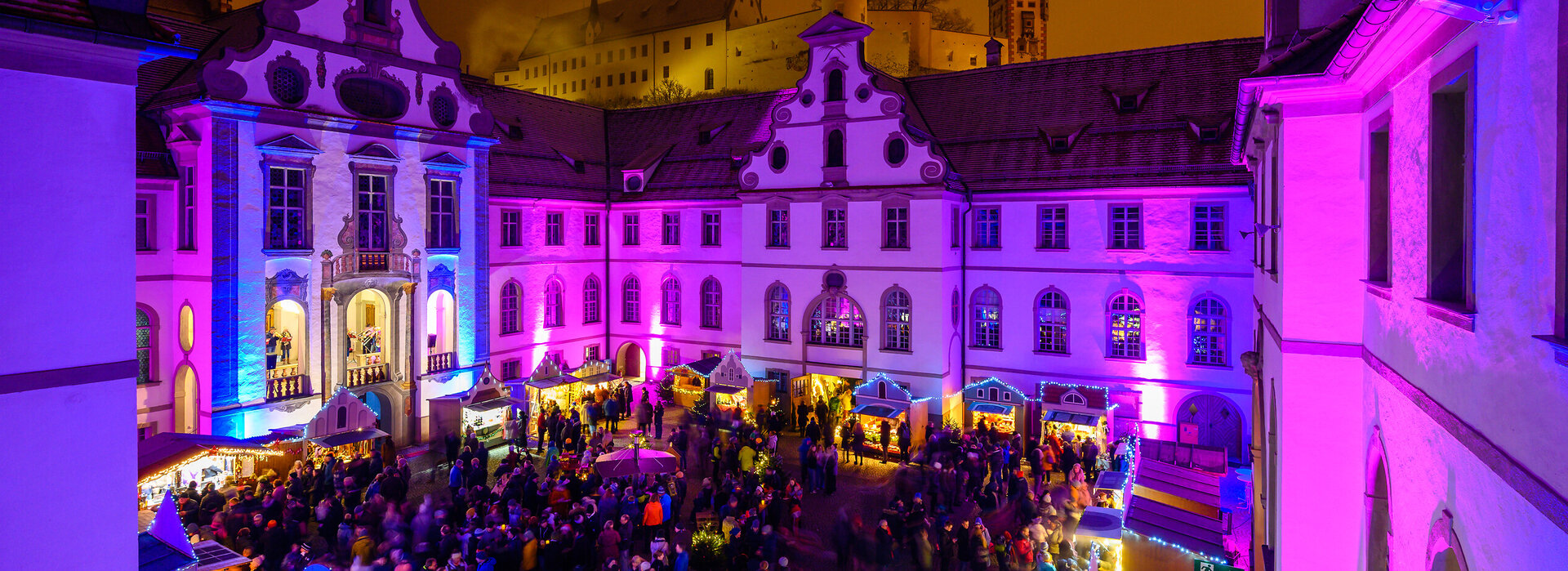 Stimmungsvoll beleuchteter Adventsmarkt mit vielen Menschen im Innenhof des barocken Kloster St. Mang. 