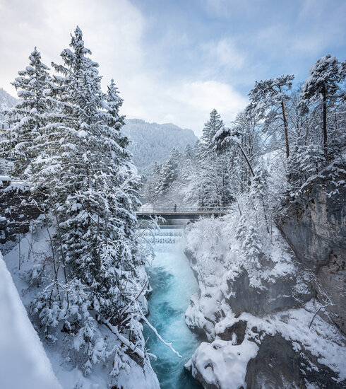 Das Bild zeigt einen eisblauen Fluss, der sich seinen Weg durch eine Felsschlucht bahnt. Über den Fluss führt eine Brücke. Die Felsen und die weitere Umgebung sind verschneit. Der Himmel ist blau.   