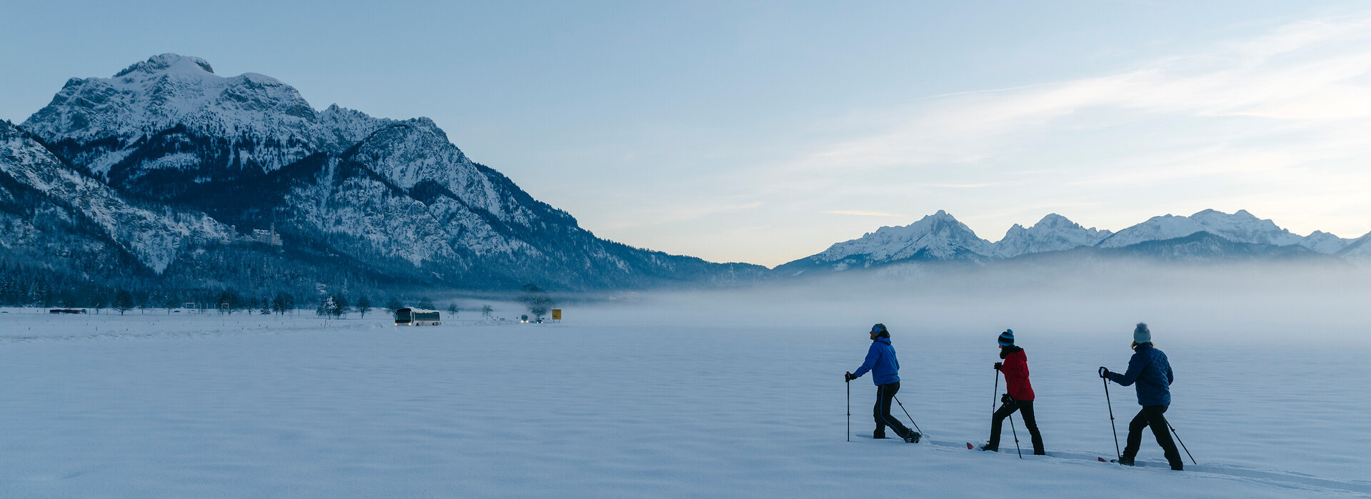 Das Bild zeigt eine flache, schneebedeckte Ebene, über die drei Schneeschuhwanderer hintereinander laufen. Leichte Nebelschwaden hängen über dem Boden. Im Hintergrund erhebt sich der mächtige Gipfel des Säulings.