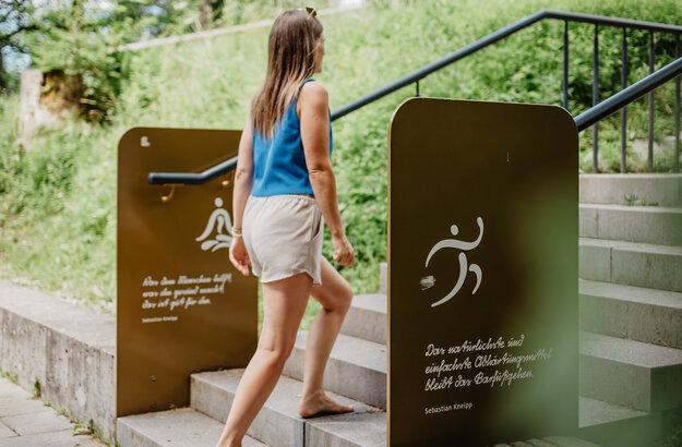 Das Bild zeigt eine Frau, die eine Treppe zwischen zwei Kneipp-Tafeln hinauf geht