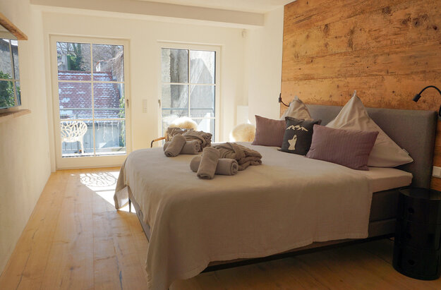 Ein gemütliches Schlafzimmer mit Blick durch die Flügeltüren. Die Wände sind mit Holzpaneelen verkleidet, die Einrichtung ist schlicht und modern. 