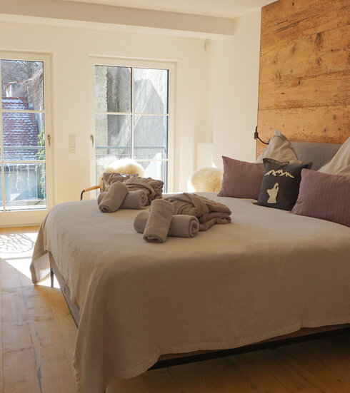 Ein gemütliches Schlafzimmer mit Blick durch die Flügeltüren. Die Wände sind mit Holzpaneelen verkleidet, die Einrichtung ist schlicht und modern. 