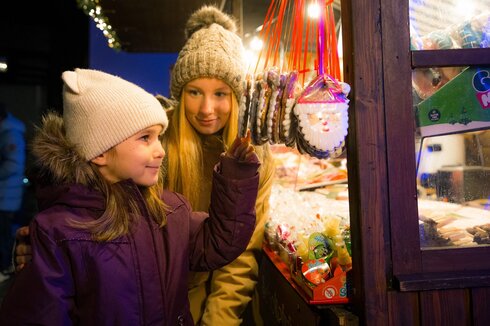Auf dem Bild sieht man ein jüngeres und ein älteres Mädchen die gemeinsam an einem Süßigkeitenstand Lebkuchen-Weihnachtsmänner betrachten.