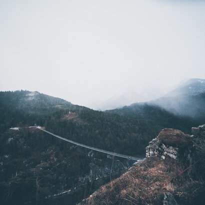 Das Bild zeigt eine Hängebrücke, die zwei Bergeplateaus miteinander verbindet. Auf der einen Anhöhe ist eine Burgruine zu sehen. Die Berge sind mit Wald bewachsen. Der Himmel ist rau. Nebel zieht auf. 