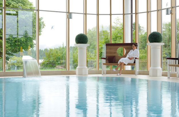 Das Bild zeigt eine Frau, die auf einer edlen Holzbank neben einem Indoorpool sitzt. Sie trägt einen weißen Bademantel. Durch die große Fensterfront im Hintergrund sind grüne Büsche und Bäume zu sehen. Der Himmel ist grau.  