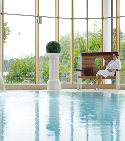 Das Bild zeigt eine Frau, die auf einer edlen Holzbank neben einem Indoorpool sitzt. Sie trägt einen weißen Bademantel. Durch die große Fensterfront im Hintergrund sind grüne Büsche und Bäume zu sehen. Der Himmel ist grau.  
