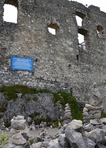 Das Bild zeigt eine majestätische Burg ähnliche Struktur, erbaut aus grauem Stein. Die Burg zeichnet sich durch ihre vielen Fenster und ein großes, blaues Schild aus, das auffällig an der Seitenfront des Gebäudes angebracht ist. Die Burg liegt auf einem Hügel, umgeben von einer üppigen grünen Landschaft. Der Himmel über ihr ist ein klarer blauer Himmel, der die ganze Szene ästhetisch ansprechend gestaltet. Die Größe und Schönheit der Burg und die friedliche Umgebung machen einen faszinierenden Anblick aus.