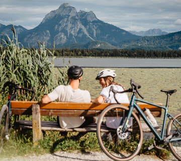 Das Bild zeigt eine junge Frau und einen Mann, die in Fahrradkleidung auf einer Bank am Seeufer sitzen. Ein Fahrrad lehnt an der Bank. Im Hintergrund erheben sich mächtige Berge.