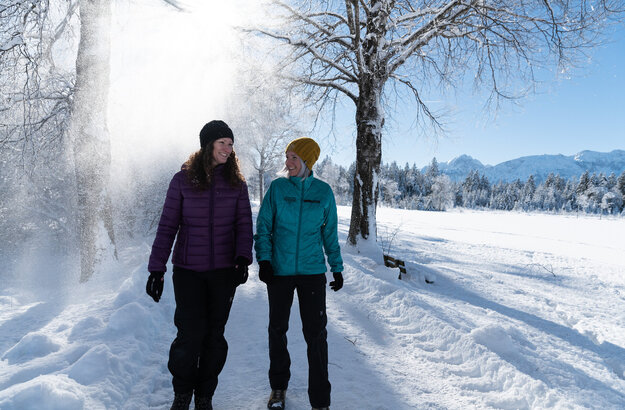 Das Bild zeigt zwei Frauen, die in warmer Winterkleidung auf einem geräumten Weg durch eine glitzernde Winterlandschaft laufen. Der Schnee rieselt von den Bäumen und im Hintergrund sind weiße Gipfel der Berge zu erkennen.