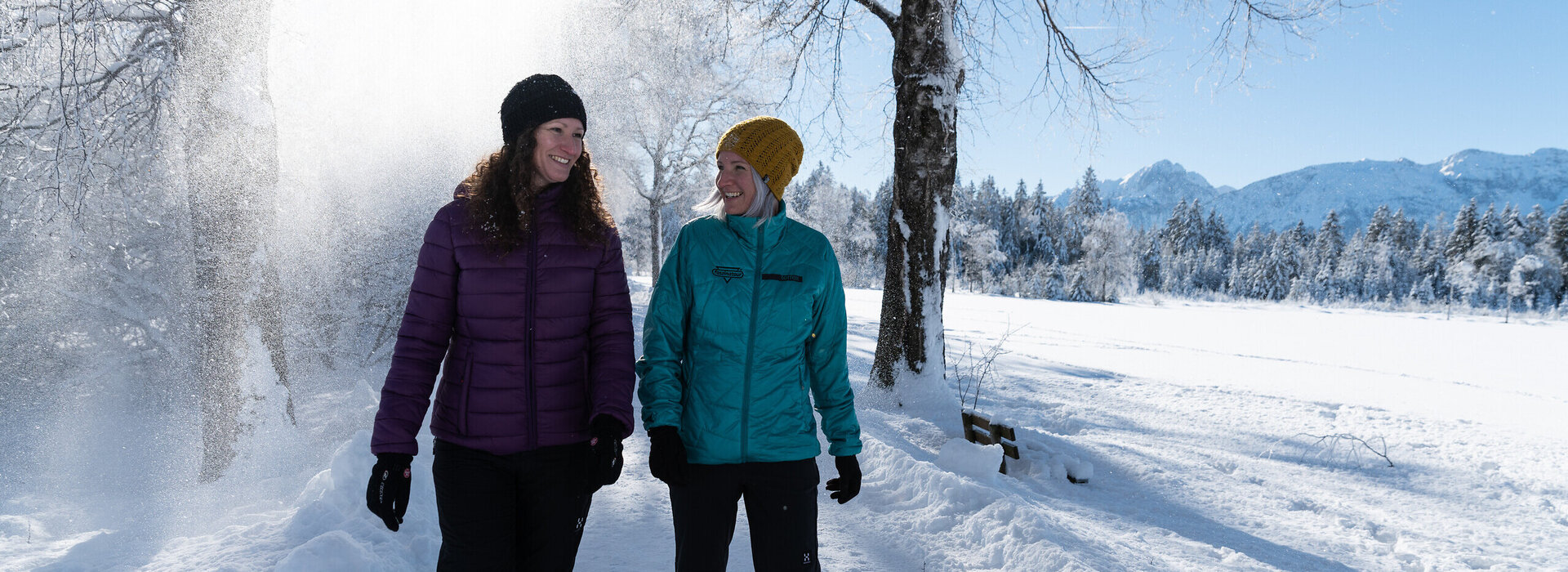 Das Bild zeigt zwei Frauen, die in warmer Winterkleidung auf einem geräumten Weg durch eine glitzernde Winterlandschaft laufen. Der Schnee rieselt von den Bäumen und im Hintergrund sind weiße Gipfel der Berge zu erkennen.