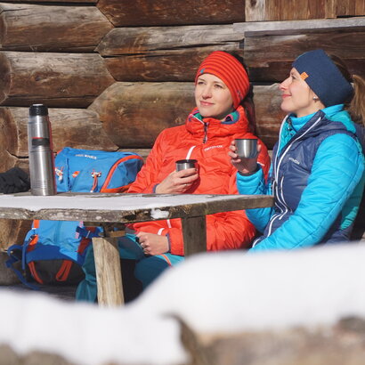 Das Bild zeigt zwei Frauen, die an einem Holztisch vor einem Holzstadel sitzen. Nur ein Detail der Wand ist zu sehen. Beide tragen Winterkleidung und Stirnbänder. Sie haben je einen Thermoskannenbecher in der Hand. Die Thermoskannen stehen auf dem Tisch. Die Sonne scheint. Ringsum ist alles schneebedeckt. 