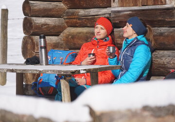 Das Bild zeigt zwei Frauen, die an einem Holztisch vor einem Holzstadel sitzen. Nur ein Detail der Wand ist zu sehen. Beide tragen Winterkleidung und Stirnbänder. Sie haben je einen Thermoskannenbecher in der Hand. Die Thermoskannen stehen auf dem Tisch. Die Sonne scheint. Ringsum ist alles schneebedeckt. 