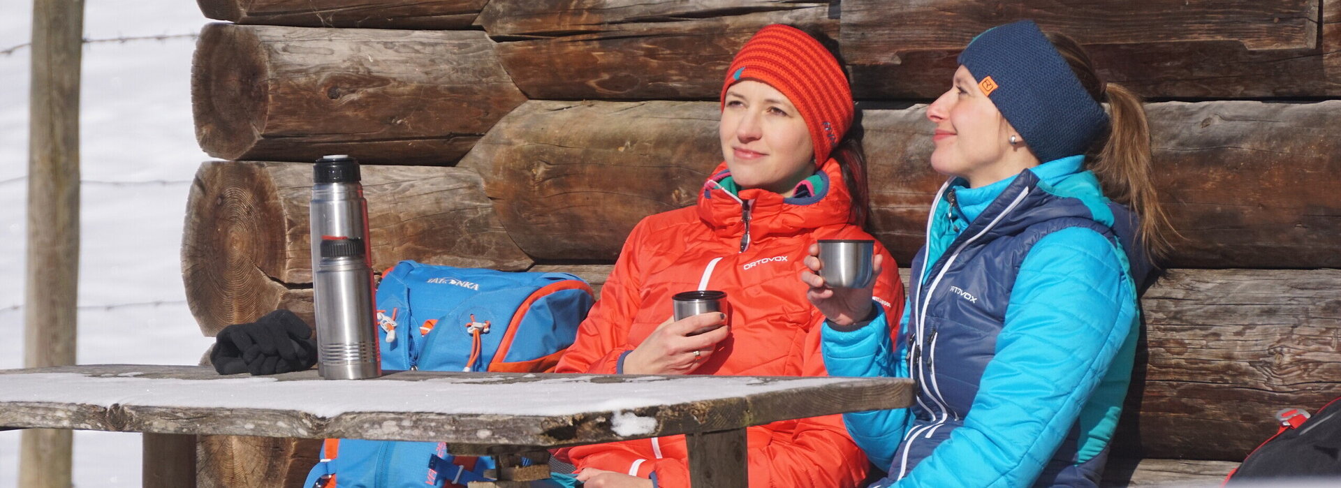 Das Bild zeigt zwei Frauen, die an einem Holztisch vor einem Holzstadel sitzen. Nur ein Detail der Wand ist zu sehen. Beide tragen Winterkleidung und Stirnbänder. Sie haben je einen Thermoskannenbecher in der Hand. Die Thermoskannen stehen auf dem Tisch. Die Sonne scheint. Ringsum ist alles schneebedeckt. 