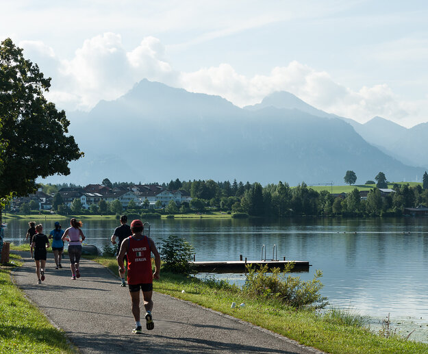 Das Bild zeigt einen See im Bergen im Hintergrund bei schönem Wetter. An der Uferpromenade des Sees joggen sportlich angezogene Menschen.