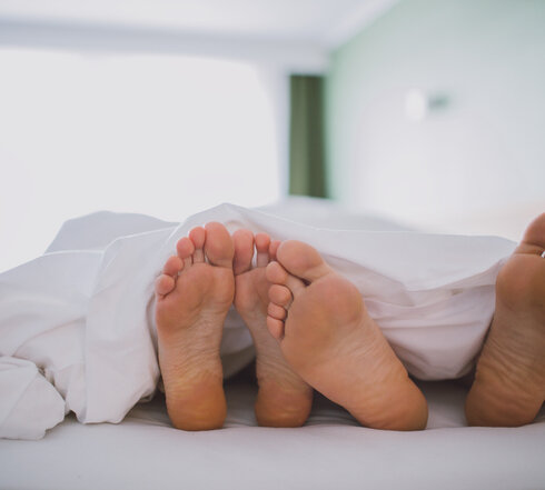 Das Bild zeigt zwei nackte Fußpaare, die unter einer weißen Decke hervorschauen. Die Füße sind so positioniert, dass sie fast verborgen sind. Die Decke, die das Hauptmotiv des Bildes ist, ist ordentlich verstaut, was eine behagliche und einladende Atmosphäre schafft. Die Kulisse ist verschwommen, was die Hauptmotive - die Füße und die Decke - betont. Die Gesamtzusammenstellung des Bildes suggeriert einen Moment der Entspannung und des Wohlbehagens