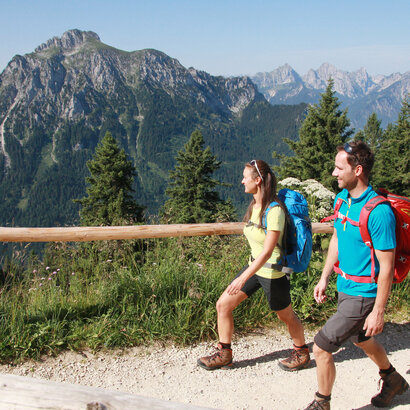 Das Bild zeigt einen Mann und eine Frau in kurzer Wanderkleidung in auf einem unbefestigten Weg laufen. Parallel zum Kiesweg verläuft ein Holzgeländern. Im Hintergrund erheben sich die Berge. Der Himmel ist blau. Die Sonne scheint. 