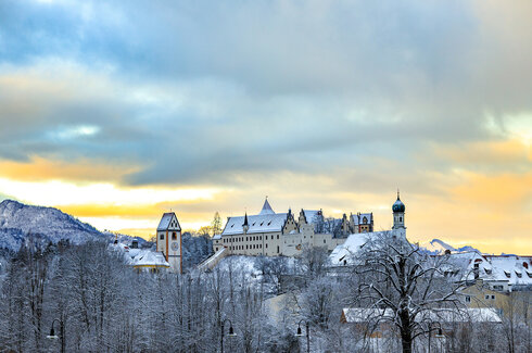 Winterliche Ansicht der Füssener Altstadt mit schneebedeckten Dächern. Um die Altstadt herum sind schneebedeckte Bäume und ein bunter Himmel zu sehen.