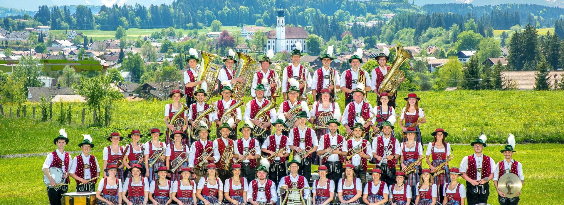 Vereinsbild einer Musikkapelle im Frühling im Ostallgäu. Im Hintergrund ist viel natur zu sehen und ein Kirchturm sticht im Hintergrund heraus.
