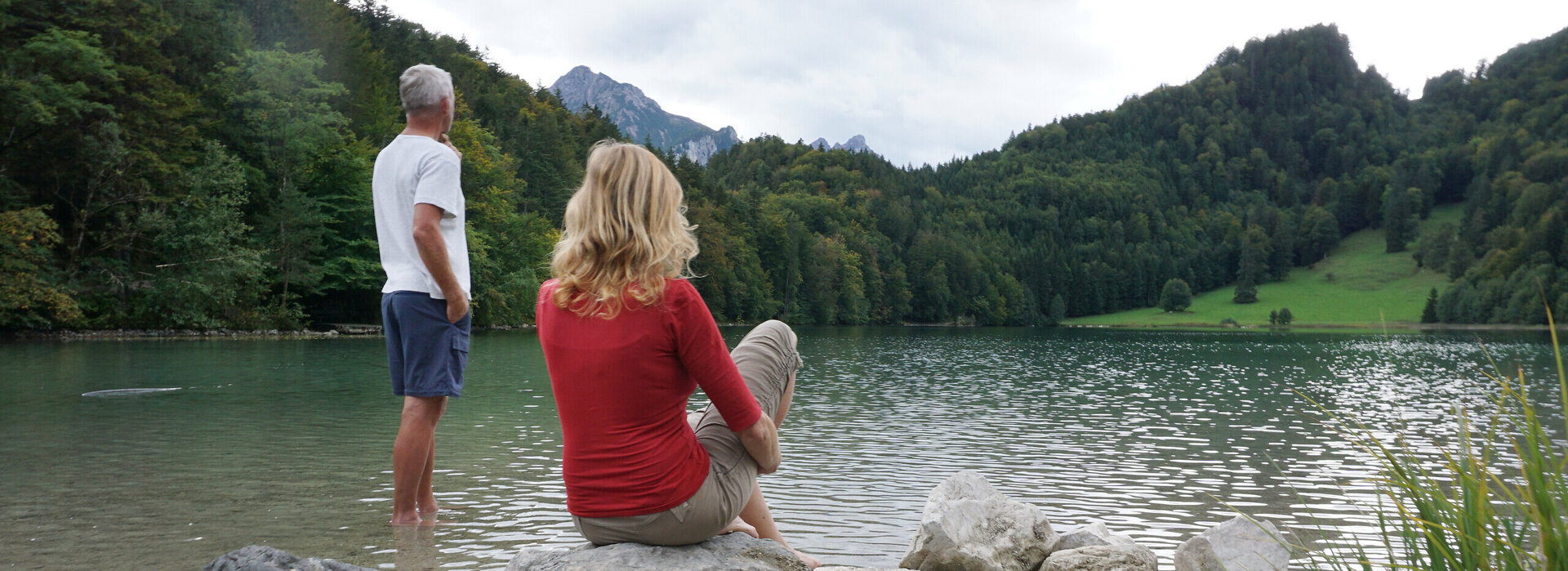 Man blickt auf eine Frau und einen Mann die auf einen See blicken. Die Frau sitzt auf einem großen Stein am Ufer. Der Mann steht im köcheltiefen, ufernahen Wasser. Der See ist ringsum mit grünen Wäldern umgeben. Dahinter erheben sich die Berge. Der Himmel ist wolkenverhangen. 
