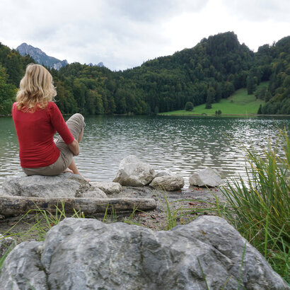 Man blickt auf eine Frau und einen Mann die auf einen See blicken. Die Frau sitzt auf einem großen Stein am Ufer. Der Mann steht im köcheltiefen, ufernahen Wasser. Der See ist ringsum mit grünen Wäldern umgeben. Dahinter erheben sich die Berge. Der Himmel ist wolkenverhangen. 