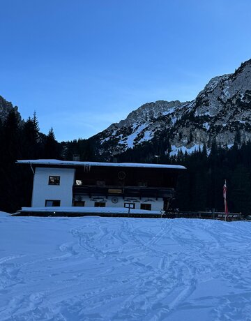 Schneefeld mit Spuren vor der Berghüttte Vilser Alm mit weißer Fassade und dunklem Obergeschoss. Zwei Flaggen, Nadelwald und schneebedeckte Berge unter blauem Himmel.