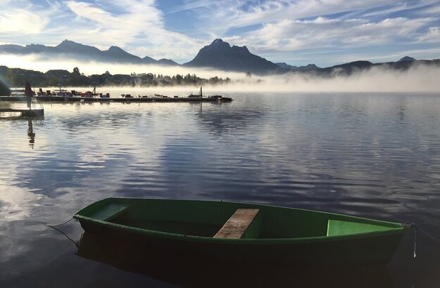 Das Bild fängt eine friedliche Szene eines kleinen grünen Bootes ein, das sanft auf dem Hopfensee schwimmt. Das Boot befindet sich im Vordergrund, hinter ihm breitet sich der weite Horizont des Wassers aus. Im Hintergrund erhebt sich die majestätische Bergkette der Ammergauer Alpen, die teilweise von einer Schicht Nebel verdeckt sind. Der Himmel über ist ein klares Blau, durchsetzt mit einigen Wolken. 