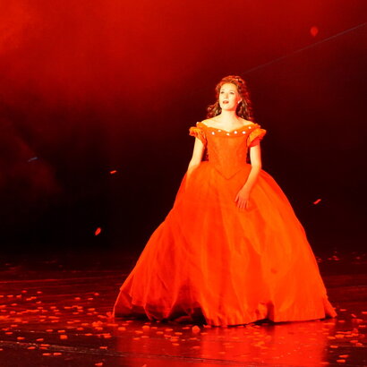 Das Foto zeigt eine Frau in einem historischen roten Kleid mit einem sehr weiten ausladenden, roten Rock, der bis zum Boden reicht. Die Frau hat lange braune Haare. Die Scene ist in rotes Licht getaucht. Am Boden liegen unzählige rote Rosenblätter. Die Szene zeigt die Kaiserin Sisi aus dem Musical Ludwig² - Der König ist zurück.