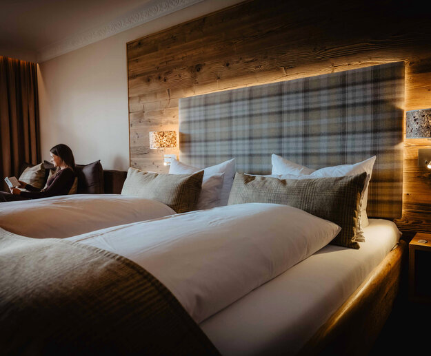 Das Bild zeigt ein Hotelbett und eine sitzende, lesende Frau im Hintergrund